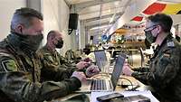 Drei Soldaten sitzen sich in einer Halle an einem Tisch gegenüber und arbeiten an ihren Laptops