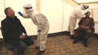Zwei Personen sitzen auf Stühlen, neben ihnen steht jeweils eine Person in Vollschutz und nimmt eine Probe aus dem Rachen