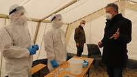 Zwei Soldaten in Vollschutz geben dem Bürgermeister und dem Generalarzt eine Notiz mit der Nummer ihrer Probe