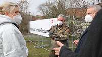 Ein Generlarzt im Gespräch mit Landrat und einer Person in Vollschutz auf einer Wiese vor dem Testzentrum