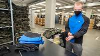 Ein junger Mann steht vor einem Tisch mit Kleidung und probiert eine Jacke an, im Hintergrund Regale und Kartons