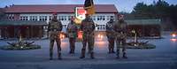 5 Soldaten stehen in der Dämmerung angetreten. Die Deutschlandflagge und ein offizielles Wappen ist zu sehen.