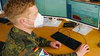 Ein Soldat in Uniform und Mund-Nasen-Schutz sitzt am Schreibtisch vor einem Computerbildschirm.
