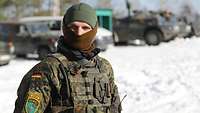Ein Soldat im Vordergrund steht auf einer schneebedeckten Straße, im Hintergrund stehen Geländefahrzeuge