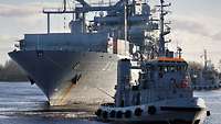 Ein großes graues Schiff, im Vordergrund ein kleiner Schlepper.