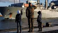 Einsatzgruppenversorger Berlin Ralf Kuchler Ein Sodat in Flecktarnuniform salutiert vor einem großen grauen Schiff im Hintergrund.