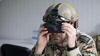 Ein Soldat trägt einen Helm. Daran ist eine Nachtsichtbrille angebracht, die er sich vor die Augen hält
