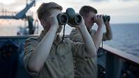 Zwei Soldaten stehen auf einem Schiff und schauen durch Ferngläser auf das offene Meer.