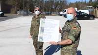Zwei Soldaten stehen am neuen Standort und halten eine Urkunde.