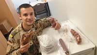 Ein Soldat mit einer Wurst und einem Schinken in der Hand lehnt am Kühlschrank. Darauf liegen verschiede Wurstsorten
