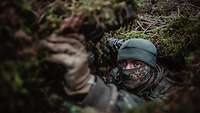 Ein Soldat befindet sich gut getarnt im Versteck. Nur sein oberes Gesichtsfeld macht ihn erkennbar.