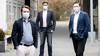 Drei Männer mit Mund- und Nasenschutzmaske stehen mit Abstand zueinander vor einem Gebäude