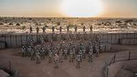 Soldatinnen und Soldaten stehen angetreten in einem Camp in der Wüste, im Hintergrund geht die Sonne unter