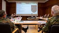 In einem Tagungssaal sitzen mehrere Soldaten an Tischen, die zu einem U aufgestellt sind. Sie blicken auf eine große Leinwand.