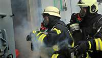 Zwei Soldaten mit Atemmasken und Schutzhelmen in schwarz-gelben Feuerwehranzügen schauen in einem verqualmten Raum.