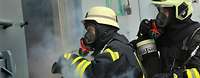 Zwei Soldaten mit Atemmasken und Schutzhelmen in schwarz-gelben Feuerwehranzügen schauen in einem verqualmten Raum.