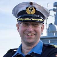 Ein lächelnder Marineoffizier in dunkelblauer Arbeitsuniform und weißer Schirmmütze.