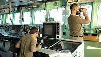 Zwei Soldaten in sandfarbener Marineuniformarbeiten stehen auf einer Schiffbrücke; einer blickt durch ein Fernglas.
