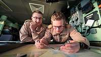Zwei Soldaten in sandfarbener Marineuniform schauen auf eine Seekarte.