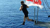 Ein Soldat im schwarzen Schwimmanzug springt mit einer rot-weißen Flagge ins Wasser.