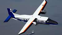 Ein zweimotoriges Propellerflugzeug über dem Meer. Darunter ein Tanker.