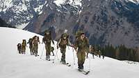 Vor einem großen Berg in den Alpen wandert eine Gruppe Soldaten mit Tourenskiern schräg bergauf.