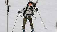 Ein Soldat auf Skiern mit Schneetarnjacke und Rucksack fährt einen Hang hinunter.