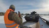 Ein Soldat mit einer Warnweste in Signalfarben steht vor einem auf ihn zufahrenden Panzer