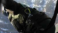 Ein Soldat in grünem Fliegeranzug blickt im Flug aus der Seitentür eines Hubschraubers.
