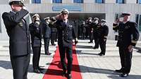 Ein Admiral in dunkelblauer Marineuniform und weißer Schirmmütze geht grüßend einen roten Teppich entlang.