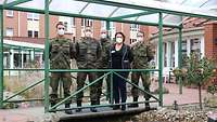 Vier Soldaten mit Flecktarnuniform und eine Frau in Zivil stehen mit hinter dem Rücken verschränkten Händen vor einem Haus.