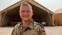 Ein Soldat lächelt in die Kamera. Im Hintergrund stehen zwei Zelthallen, in einer davon steht ein Hubschrauber