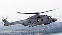 Ein Hubschrauber fliegt, im Hintergrund sieht man Wasser, Gebäude und Wald