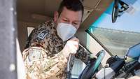 Ein Soldat mit Mund-Nasen-Schutz sitzt in einem Auto und hält sich das Funkgerät an den Mund