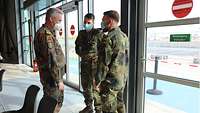 Ein Generalarzt unterhält sich mit zwei Soldaten in einem Flughafengebäude. Im Hingergrund ist der Außenbereich zu sehen.