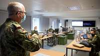 Ein Soldat steht vor mehreren Soldatinnen und Soldaten, die an Tischen sitzen. Alle tragen Mund-Nasen-Schutz