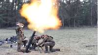 Zwei Soldaten hocken neben einem Mörser, der einen Schuss mit Feuerstrahl abgibt. Ein Soldat hält sich stehend die Ohren zu.