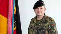 Ein Bundeswehrgeneral in Flecktarnuniform hat ein schwarzes Barett auf. Er steht neben einer schwarz-rot-goldenen Fahne.