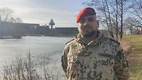 Ein Soldat steht neben einem See und lächelt in die Kamera. Im Hintergrund stehen mehrere Bäume und Gebäude