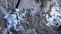 Soldaten in Schneetarnanzügen Rucksäcken und Skiausrüstung steigen einen sehr schmalen Pfad an einer Felswand entlang.