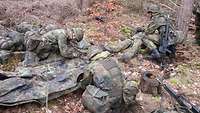 Soldaten bauen eine behelfsmäßige Trage. Zwei Soldaten bauen aus Zeltplanen und Ästen eine Trage. Ein dritter Soldat hockt bei dem Verwundeten, der liegt.
