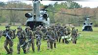 Auf einer Wiese stehen zwei militärische Transporthubschrauber, aus denen Soldaten mit Waffen und Ausrüstung kommen.