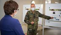 Ein Soldat weist mit einem Zeigestock auf eine Infotafel und spricht dabei mit einer Zivilistin, die vor ihm steht.