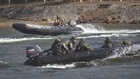 Auf dem Wasser kreuzen sich zwei Schlauchboote mit Soldaten, Gischt hinter den Außenbordmotoren.