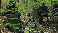 Ein verwundeter Soldat lehnt im Wald an einem Baum, drei Kameraden kümmern sich um ihn.