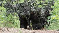 Mehrere Soldaten laufen unter Tarnnetzen im Wald entlang, die vorderen haben Baumstämme bei sich.