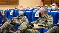 Soldaten mit Mund-Nasenschutz sitzen in einem Vortragssaal auf blauen, gepolsterten Klappsesseln.