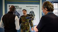 Ein Soldat steht vor einer Kamera in einem Büroraum und spricht mit einem Zivilisten.