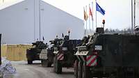 Zwei Schützenpanzer Marder und zwei Transportpanzer Fuchs fahren hintereinander in die Kaserne. Im Hintergrund wehen Flaggen