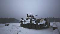 Ein Schützenpanzer steht in Stellung. Auf ihm steht ein Soldat. Im Hintergrund steht eine Gruppe von Personen 